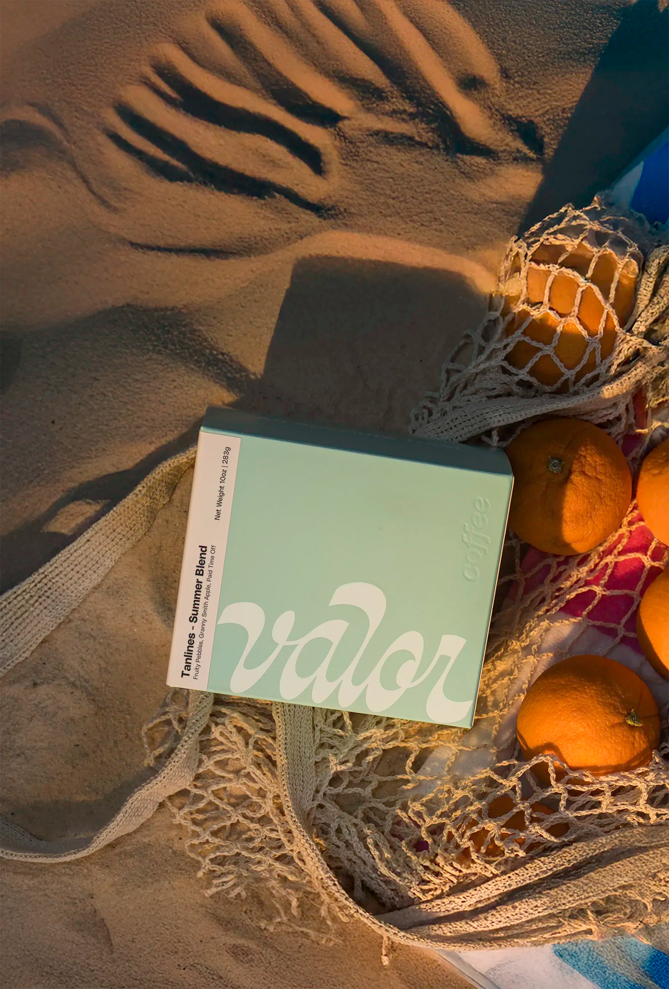 A teal box of Valor's Tanlines Summer Blend sits on a beach under warm, setting sunlight. The box is next to a beach towel and a cotton mesh bag of citrus.